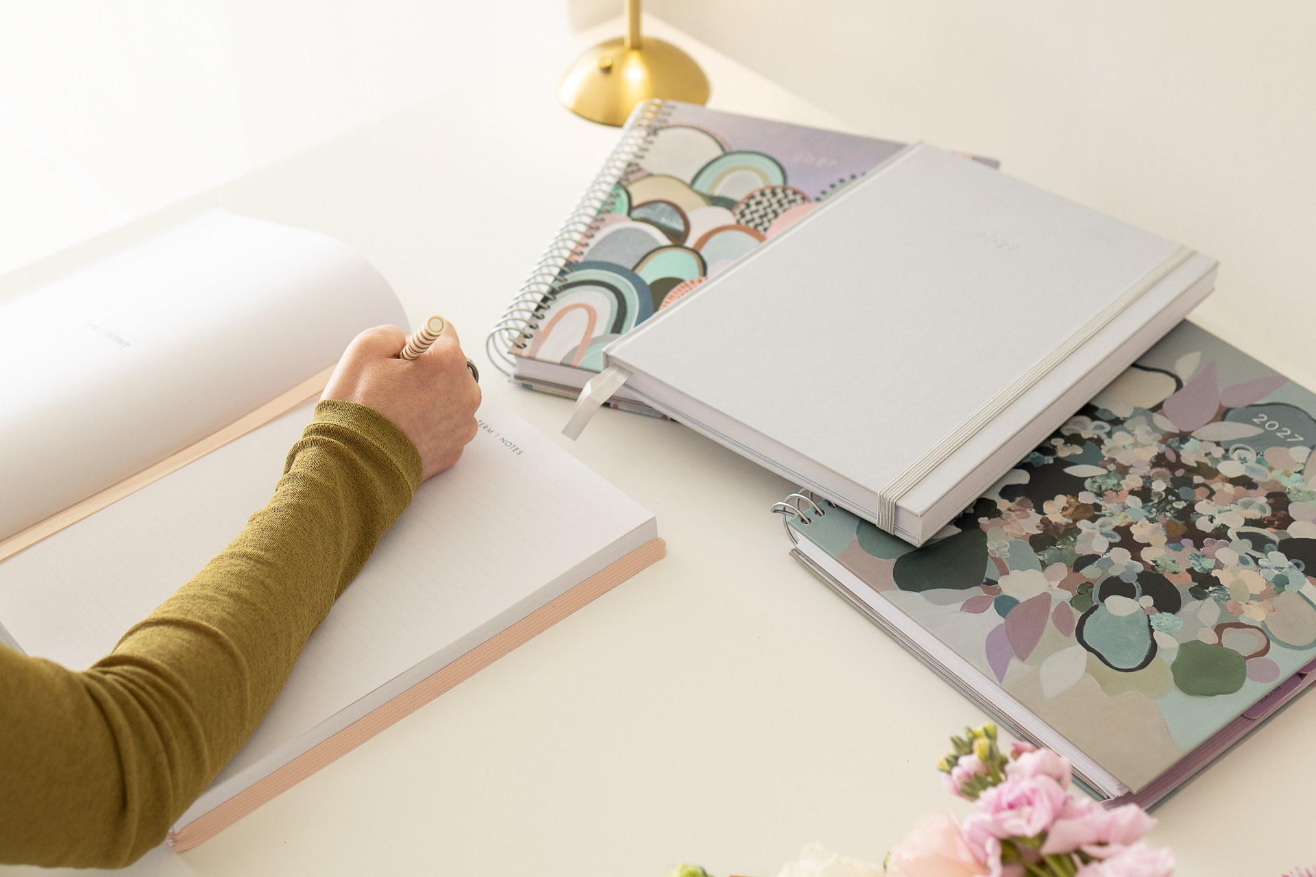 A hand writing in the 2027 planner, with the Arches, Still Life and Wildflower covers on the desk